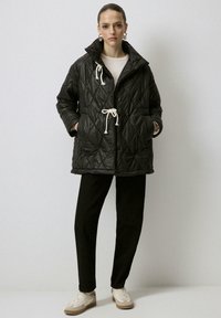 Chaqueta negra acolchada con un alto cuello y cordón crema, que presenta dos bolsillos delanteros y un cierre de botones. Llevada sobre pantalones negros y zapatillas.