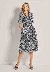 Schwarzes und weißes, gemustertes Kleid mit V-Ausschnitt, kurzen Ärmeln und taillierten Bund. Das Material scheint leicht zu sein; knielang mit Taschendetails.