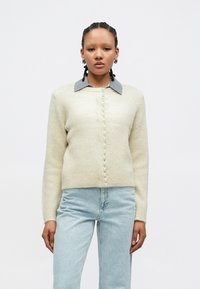 Cardigã creme com padrão canelado e gola cinzenta, com fecho frontal de botões semelhantes a pérolas. Combinado com jeans azul claro.