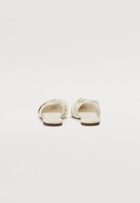 White slide-on sandals made of soft material, featuring a knotted design at the front, flat sole, and minimal stitching details.