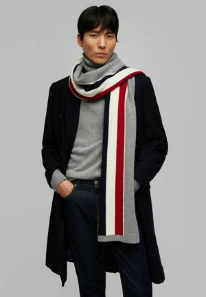 Écharpe tricotée avec des rayures grises, blanches, rouges et bleu marine. Texture douce. Portée avec un col roulé gris et un jean foncé, superposé sous un manteau bleu marine.