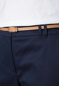 Pantalon marine avec une ceinture en cuir beige et des accessoires en argent. La ceinture est enfilée dans les passants et se situe à la taille au-dessus d'une chemise blanche.