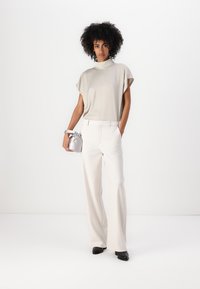 Lichtbeige top met korte mouwen, brede witte broek en zwarte schoenen. Model houdt een kleine zilveren handtas vast. Eenvoudig, modern ontwerp.
