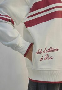 Bílá mikina s červenými pruhy na ramenou a vyšitým textem "club d’athlétisme de Paris," s žebrovanými manžetami a spodním lemem.