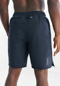 Granatowe spodenki sportowe z elastycznym paskiem, zapinaną na zamek tylną kieszenią oraz odblaskowymi akcentami po bokach. Gładki materiał o lekkiej teksturze.