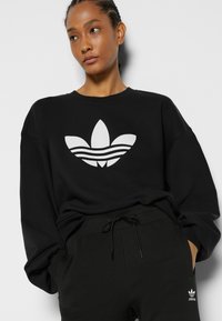 Czarny sweter z dużym białym logo trefoil z przodu. Posiada luźny krój, prążkowany dekolt oraz długie rękawy. W zestawie z czarnymi spodniami.