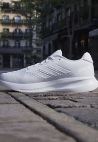 Chaussure de course légère blanche avec empeigne en mesh et semelle texturée sur une rue pavée avec un arrière-plan de bâtiments flous.