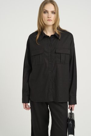Svart skjorte med knapper, lange ermer, to brystlommer og en avslappet passform. Stoffet har en teksturert overflate. Sammen med svarte bukser.