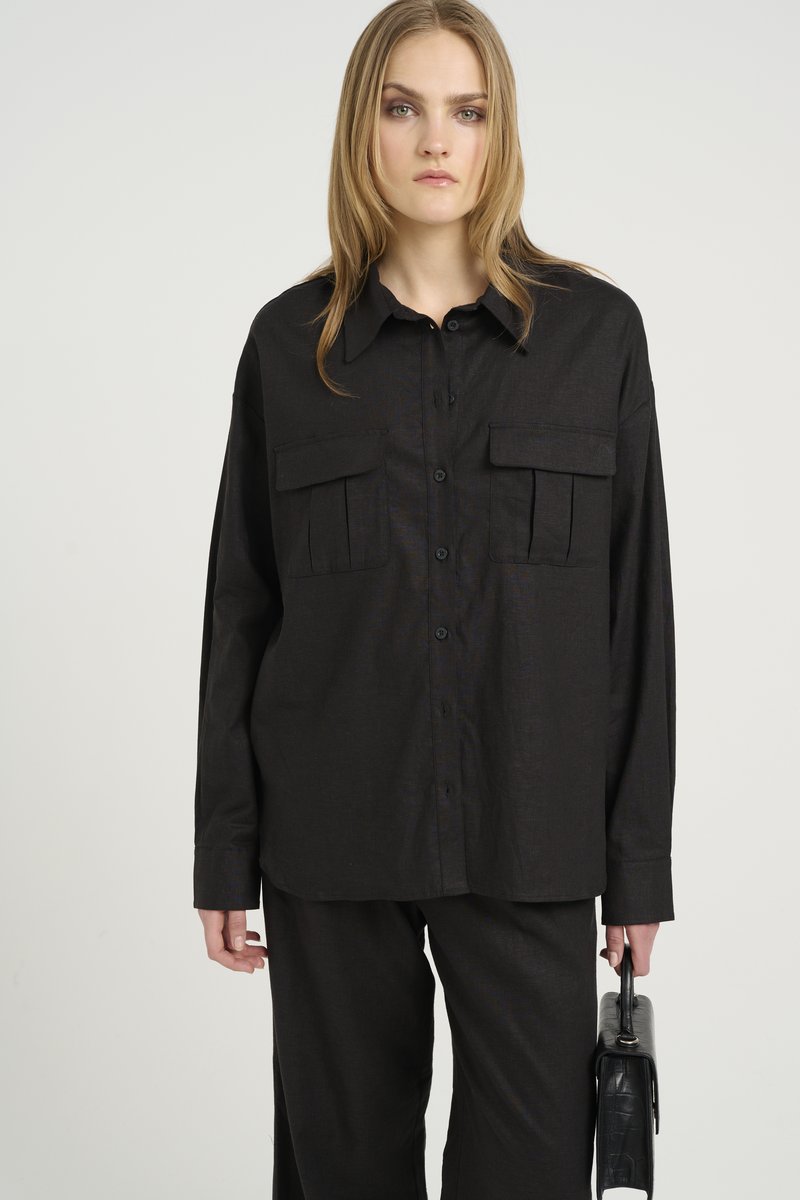 Camicia nera con bottoni, maniche lunghe, due tasche sul petto e vestibilità rilassata. Il tessuto ha un aspetto texture. Abbinata a pantaloni neri.