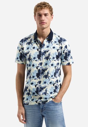 Jonge man die een blauw-wit gebloemd poloshirt met korte mouwen en een donkere kraag draagt, gecombineerd met lichtblauwe jeans, staand met één hand in zijn broekzak.