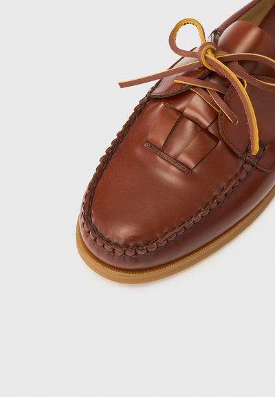 Καφέ δερμάτινο μοκασίνι με περίπλοκο υφαντό σχέδιο στο μπροστινό μέρος, κίτρινες κορδόνια που κάνουν αντίθεση και ελαφριά σόλα.