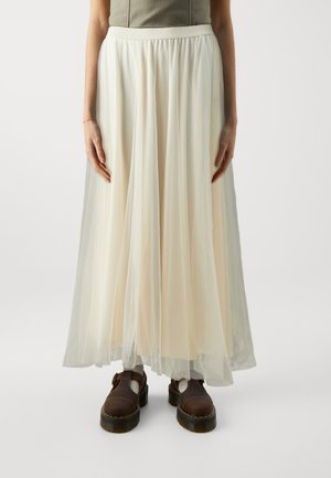 Jupe longue et fluide en tissu crémeux léger et transparent, avec une taille froncée, accompagnée de chaussures marron à semelles robustes.