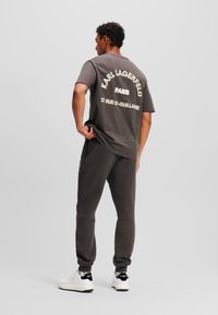 Tricou din bumbac gri cu text alb pe spate, asortat cu pantaloni de trening. Prezentând o croială lejeră și o textură moale, purtat cu sneakers albi.
