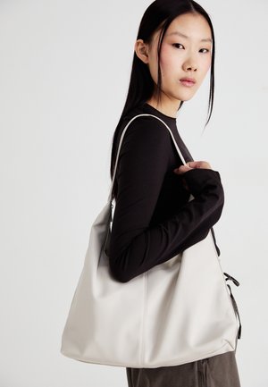 Junge Frau in schwarzem Langarmshirt trägt eine große, weiche weiße Schultertasche vor einfarbigem grauem Hintergrund.