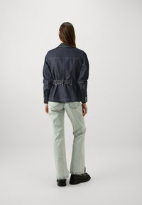 Mørk denimjakke med belte i midjen, med kragehals og knappdetaljer på ermene, kombinert med lysegrønne slengbukser.