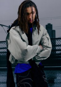 Lichtolijfgroene bomberjack met een ritszak, gedragen over een blauw-zwart gestreept shirt. Donkere broek met witte patronen.