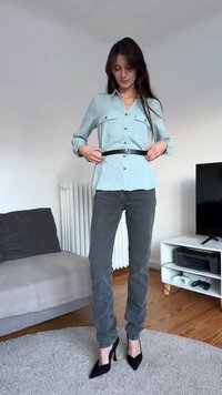 Ανοιχτό μπλε πουκάμισο με κουμπιά και τσέπες στο στήθος, μαύρη ζώνη, γκρι εφαρμοστά τζιν και μαύρες ψηλοτάκουνες γόβες με μυτερή μύτη, στέκεται σε ένα σαλόνι.