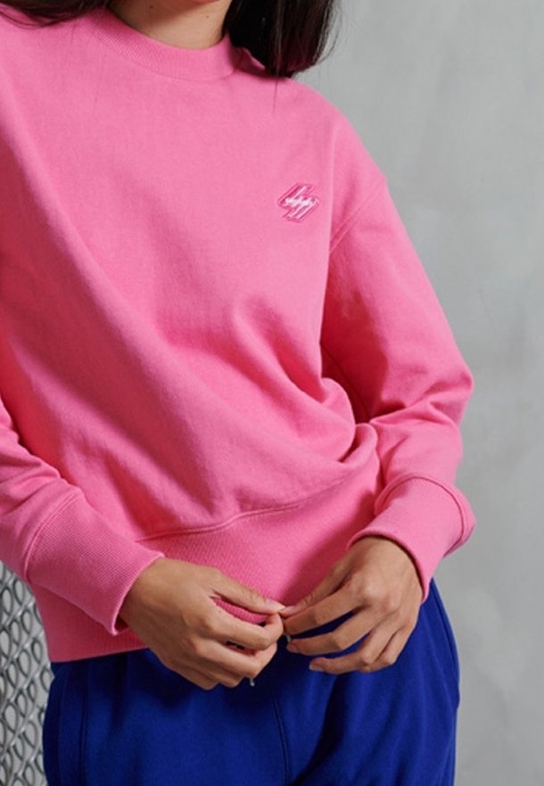 Personne portant un sweat-shirt rose vif avec un petit logo sur la poitrine et un pantalon bleu roi, ajustant l'ourlet du sweat-shirt avec les deux mains.