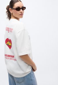 Λευκό υπερμεγέθες μπλουζάκι με κόκκινη καρδιά και κίτρινο σχέδιο αυτοκινήτου στην πλάτη. Διαθέτει κοντά μανίκια και άνετη εφαρμογή.