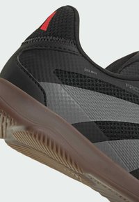 Zwarte sportieve schoen met een gestructureerde synthetische bovenkant, grijze accenten en een bruine rubberen zool met een geribbeld patroon. Rode logo op de hiel.