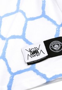 Tricou alb cu un model hexagonal albastru. Include o etichetă neagră cu logo-ul Puma și stema echipei Manchester City. Textură moale.
