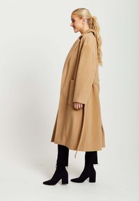 Camel wool coat with a long, straight design, oversized fit, large side pockets, and a tied waist accent. Paired with black ankle boots.