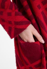 Rot gemusterter Cardigan mit einem geometrischen Design in dunklen Rottönen, weicher Textur und sichtbarem Taschen.