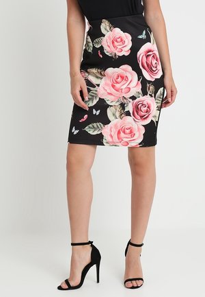 Femme portant une jupe crayon noire avec un grand motif floral rose et feuille verte, associée à des sandales noires à talons hauts avec bride à la cheville.