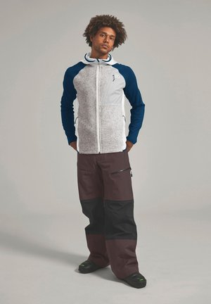 Mann mit lockigem Haar steht, trägt eine grau-navyfarbene Jacke mit Reißverschluss und braune und schwarze Hosen mit Reißverschlusstaschen vor hellem Hintergrund.