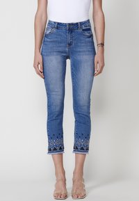 Jean en denim bleu avec une coupe ajustée, présentant des broderies décoratives à l'ourlet et des bords effilochés. Taille mi-haute et style classique avec cinq poches.