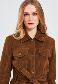 Bruine suedjacket met een kraag, voorzakken met knoopaccenten en een strikriem in de taille. Gladde textuur en een getailleerde pasvorm.