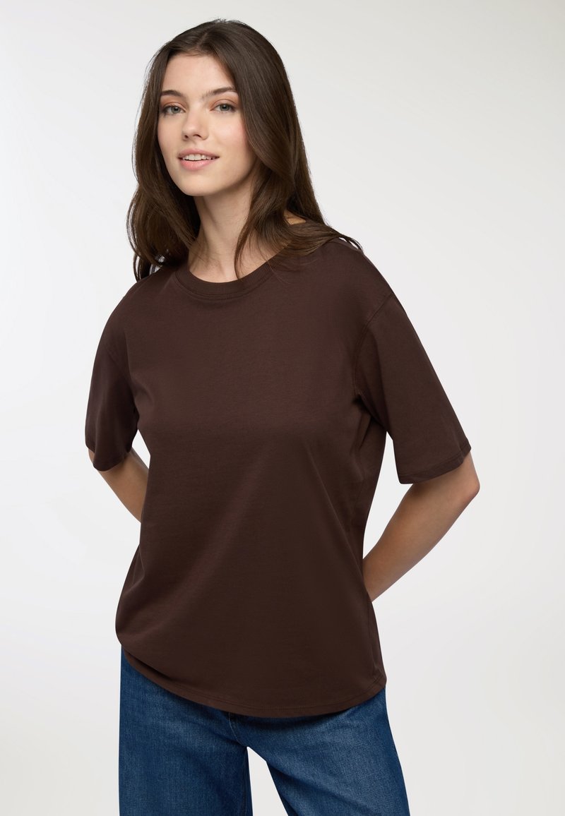 Brązowa koszulka z krótkim rękawem, wykonana z miękkiej tkaniny, z okrągłym dekoltem i luźnym krojem, w połączeniu z niebieskimi dżinsami.