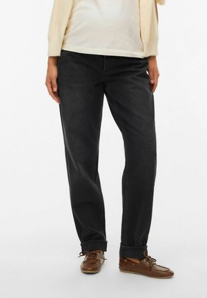 Person trägt schwarze Jeans mit geradem Bein, braune Schnürschuhe, weißes Oberteil und cremefarbener Cardigan vor einem schlichten weißen Hintergrund.