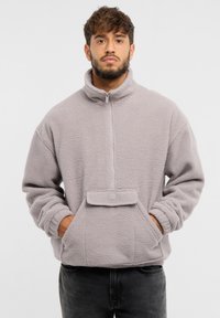 Pullover in pile grigio chiaro con colletto alto, zip frontale a metà, tasca frontale e polsini elasticizzati. Presenta una finitura morbida e strutturata.
