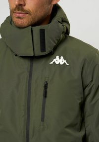 Zöld vízálló dzseki magas gallérral, fekete cipzárral és logóval a mellrészen. Simább textúrával és állítható kapucnival rendelkezik.