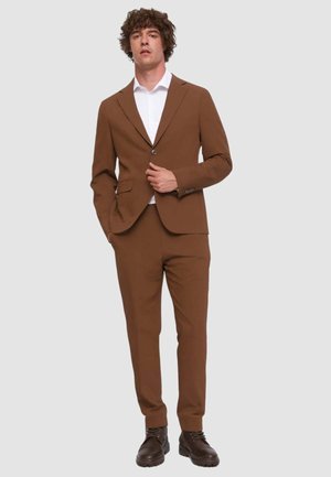 Uomo che indossa un completo marrone con camicia bianca e scarpe marrone scuro, in piedi con una mano in tasca e l'altra che sistema la giacca.