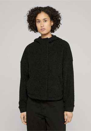 Donna con capelli ricci che indossa una giacca a cappuccio nera testurizzata e pantaloni neri, in piedi davanti a uno sfondo grigio chiaro uniforme.