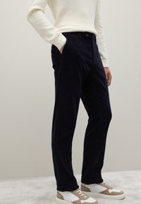 Marineblauwe corduroybroek met een rechte pasvorm en zijzakken, voorzien van een knoopsluiting. Gecombineerd met beige-witte sneakers.