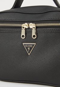 Mustakoneinen muovikäsilaukku, jossa on sileä pinta, kultainen vetoketju ja kolmion muotoinen logo, jossa lukee "GUESS" etupuolella.