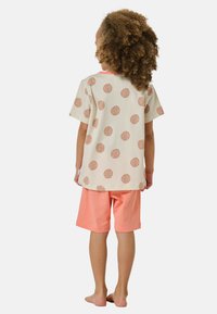 Barn med lockigt hår står barfota vänd bort, iförd en beige skjorta med bruna prickar och korallfärgade shorts.