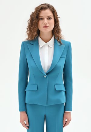 Femme aux cheveux bruns bouclés portant un costume ajusté bleu sarcelle avec un blazer à un bouton et une chemise blanche à col, debout devant un fond blanc.