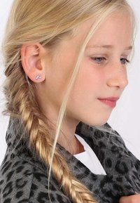 Zilveren hartvormige studoorbellen bij een jong meisje, met een gevlochten haarstijl en gekleed in een grijze dierlijke print jas.