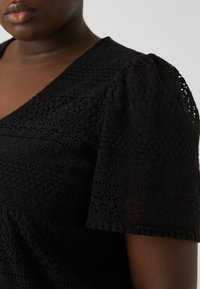 Black lace top featuring short puff sleeves, intricate patterns, textured fabric, and a V-neckline. Close-up of sleeve and shoulder area.