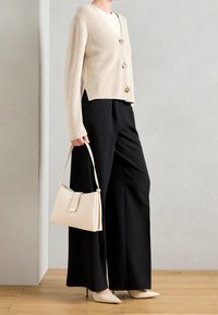 Beige gerippter Cardigan mit goldenen Knöpfen, schwarze Hose mit weitem Bein, und eine cremefarbene Handtasche. Beige spitze Absätze komplettieren das Outfit.