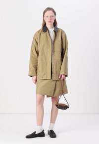Kaki regnjacka med svart krage, som matchas med en kjol i samma färg. Modellen bär svarta loafers och håller en liten brun väska. Vit bakgrund.