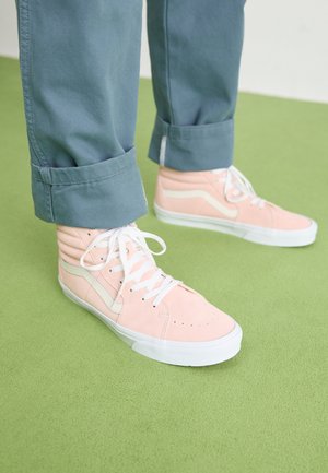 Zapatillas altas de ante rosa claro con cordones blancos y acentos laterales. Llevadas con pantalones oscuros verdes remangados sobre un fondo verde.