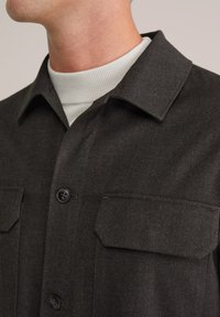 Hombre vestido con una chaqueta abotonada de color marrón oscuro con bolsillos tipo solapa sobre un suéter de cuello alto de color blanco y texturizado, con el cuello y la parte inferior de la cara visibles.
