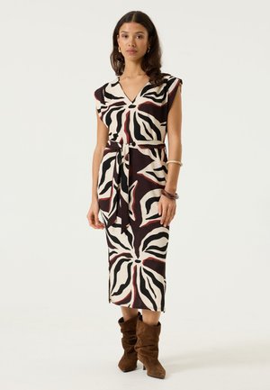Femme debout portant une robe midi sans manches à motifs avec des formes abstraites noires, crème et brunes, bottes marron souples et bracelets.