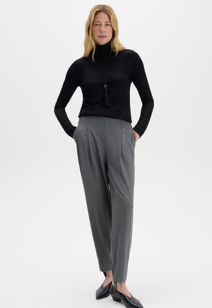 Femme aux cheveux blonds portant un col roulé noir, un pantalon gris taille haute, des chaussures noires et un long collier noir à pompons, debout les mains dans les poches.