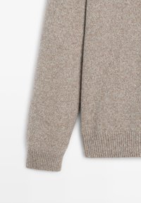 Manche de pull beige en tricot à manches longues avec détail de bord-côte sur fond blanc.